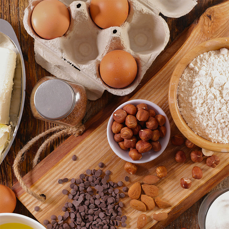 Ingredients for baking on a wooden board including eggs, flour, nuts, and chocolate chips.