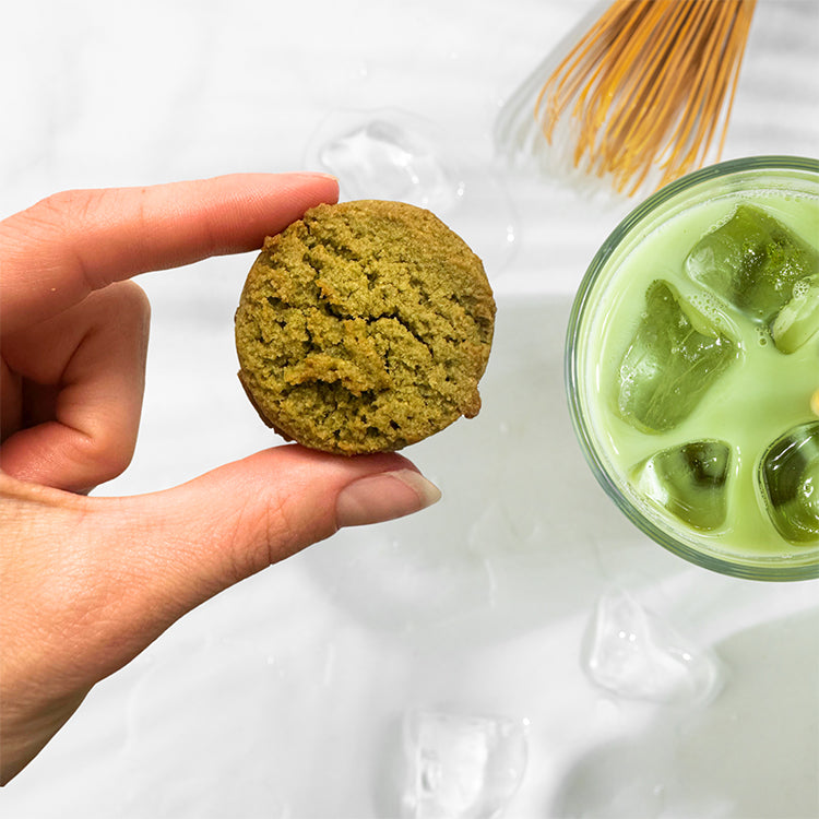 Hand holding a green matcha cookie next to a glass of matcha latte on a white background